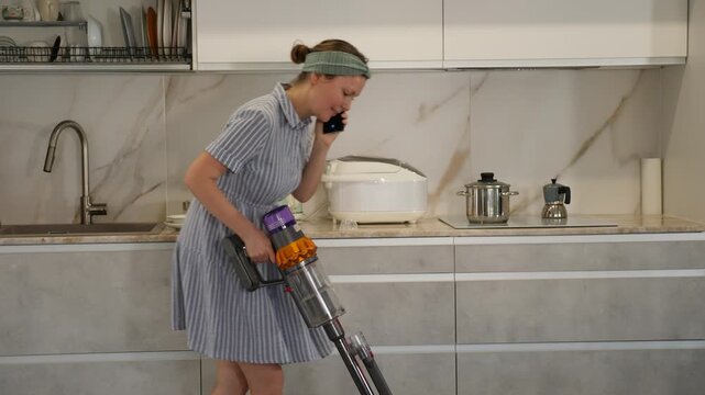 Young woman in homemade dress and headband cleans kitchen and chats on phone. Cheerful housewife is talking on mobile phone while runing vacuuming cleaning