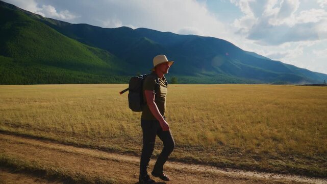 Man walks alone on dirt path through golden field. Mountains rise behind him under blue sky. He wears hat and backpack, steps confidently forward. Scene feels peaceful, adventurous, and inspiring