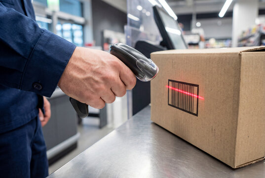 Handheld barcode scanner scanning box at retail checkout counter in a service-oriented setup. This version focuses on parcel processing at the counter with active package handling.