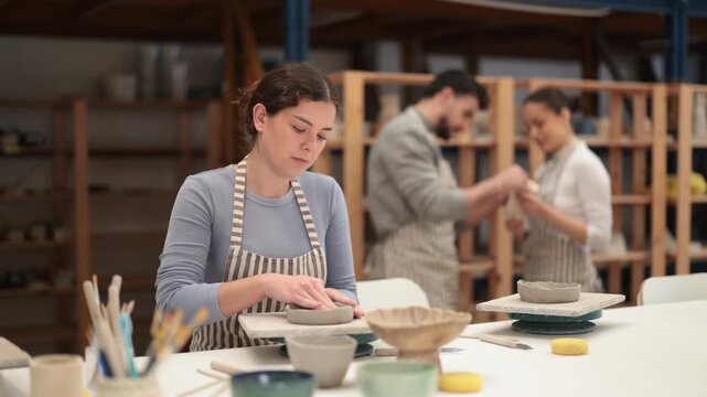 In pottery workshop, girl sitting at table, busy making products from raw clay. Amateur busy at master class modeling clay plates from clay