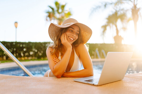 Happy woman stands in pool, working on laptop from parapet on a sunny day. A female freelancer relaxes by the pool with a laptop at sunset. Concept of remote work, relaxation, and enjoyment.