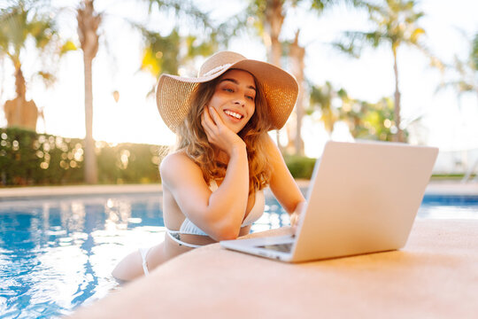 Happy woman stands in pool, working on laptop from parapet on a sunny day. A female freelancer relaxes by the pool with a laptop at sunset. Concept of remote work, relaxation, and enjoyment.