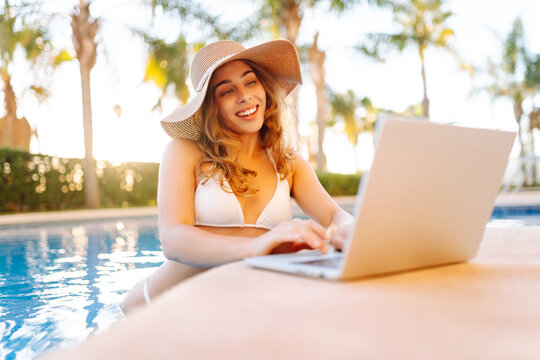 Happy woman stands in pool, working on laptop from parapet on a sunny day. A female freelancer relaxes by the pool with a laptop at sunset. Concept of remote work, relaxation, and enjoyment.