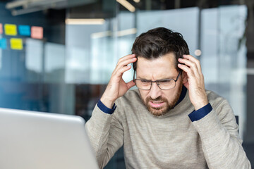 Man experiencing stress and frustration while working on laptop in a modern office, feeling the pressure of deadlines and potential burnout from professional workload © Liubomir