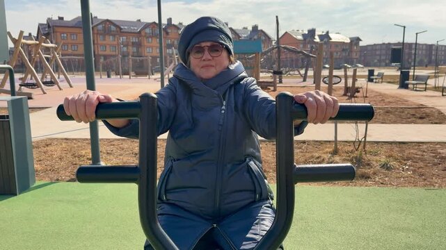 Elderly woman exercising on outdoor hand exerciser, wearing coat and cap, gripping handlebars and pedaling on green stadium turf.
