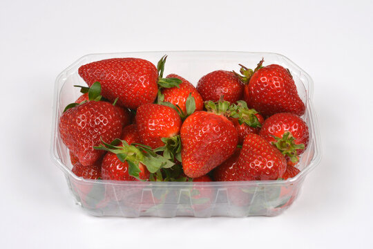 A transparent plastic punnet filled with ripe red strawberries with green leaves against a plain white background