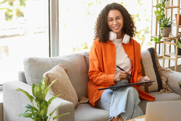 Fototapeta premium Female African-American interior designer working with graphic tablet on sofa in office