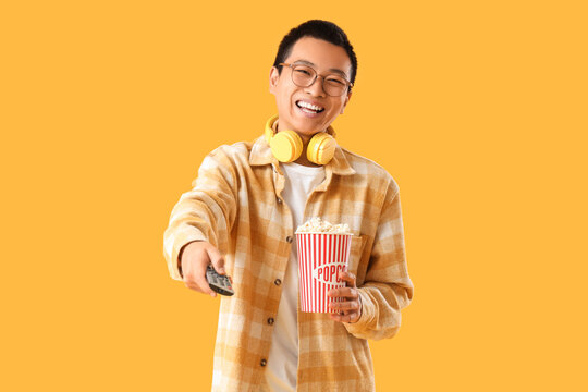 Young Asian man with TV remote controller and popcorn on yellow background