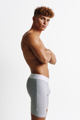 Fototapeta premium Young athletic man with curly hair posing shirtless in gray shorts against white background, showing confident expression and muscular build in studio.