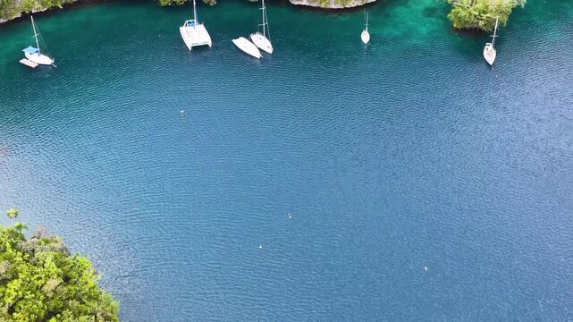 Aerial view of white sailboats and catamarans anchored in deep blue tropical lagoon surrounded by lush green limestone cliffs.