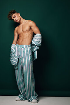 Young man, shirtless and muscular, wearing striped loose pants and loose shirt, posing on dark green background with casual posture and confident facial expression, studio fashion photo.