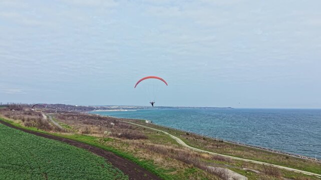A person is paragliding over the ocean near a coastline while the landscape features green fields and open water. The sky is mostly clear with some clouds visible.