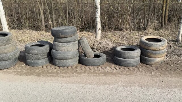 Stacked worn tires along roadside, abandoned passenger car tires piled against leafless hedgerow on spring day, gravel shoulder.