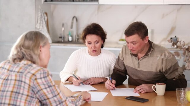 Attentive middle-aged man and woman sitting in the kitchen with sheets of paper in front of them talking to female agent