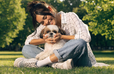 Fototapeta premium smiling young woman sitting on green grass in park, gently petting small shih tzu dog, cute fluffy dog looking at camera, summer outdoor setting, natural sunlight, blurred green trees background, warm