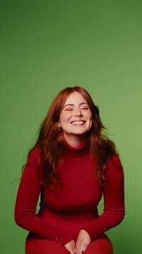 A cheerful young woman with long, red hair sits against a vibrant green backdrop, wearing a stylish red turtleneck dress, as she laughs and smiles genuinely.