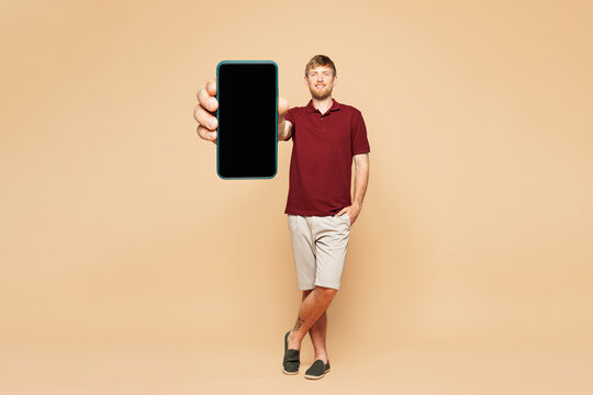 Full size young happy man wear red shirt casual clothes hold in hand use mobile cell phone with blank screen workspace area isolated on plain light beige background studio portrait. Lifestyle concept.