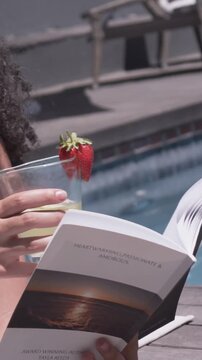 Vertical video: Reading woman in swimwear, book and glass by pool, breeze turning pages, copy space