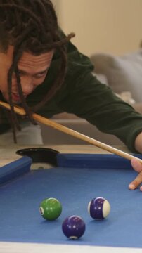 Vertical video: Leaning man in green pulling cue at home blue-felt table to sink green purple balls