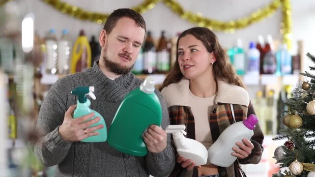 Cheerful married couple customer buying spray and detergent in small store decorated for New Year holiday 