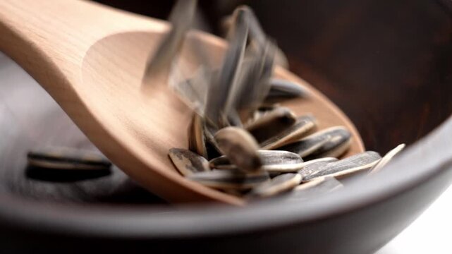 Roasted unpeeled sunflower seeds falling into a wooden spoon and a dark bowl. Healthy salty snack. Slow motion