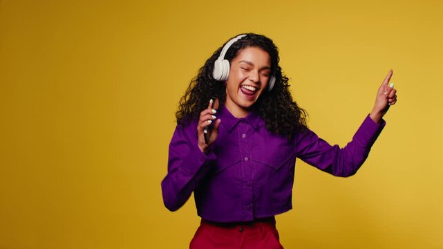 Young woman dances energetically and sings along to music on her phone while wearing headphones against a yellow background.