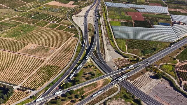 Highways intersect in a rural area with surrounding farms and greenhouses. Trucks travel on the roads while fields stretch in the background and the sun shines above.