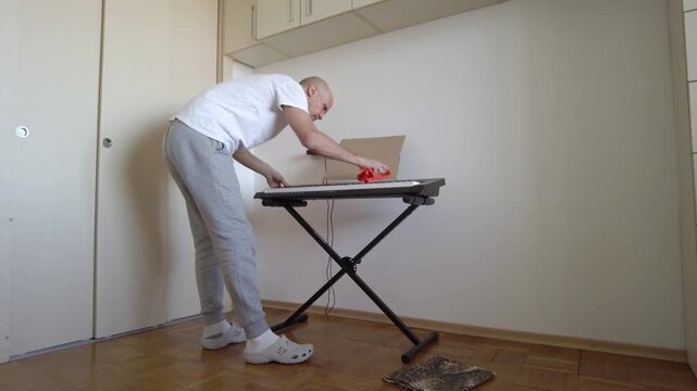Wide shot white man prepping synthesizer while standing in small apartment, wiping dust with red cloth near laptop on keyboard stand, checking cable connection and power lead,