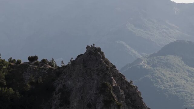 Aerial view of two people standing atop a rugged mountain peak, framed against a backdrop of hazy, layered mountain ranges, Teth, Shkoder County, Albania.