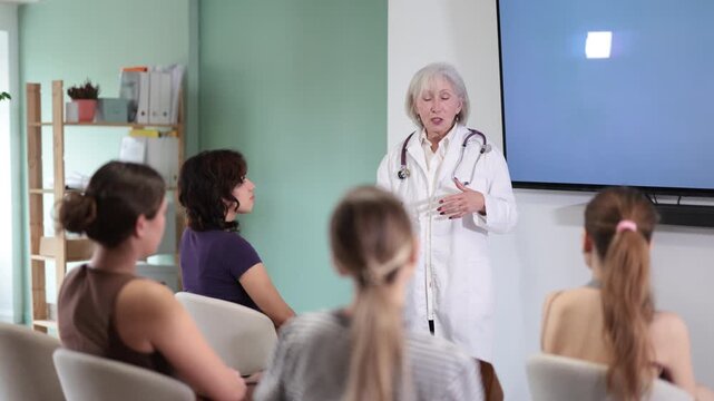 Concentrated mature female medical doctor dressed in white medical coat with stethoscope standing and talking in auditorium where sitting medical student