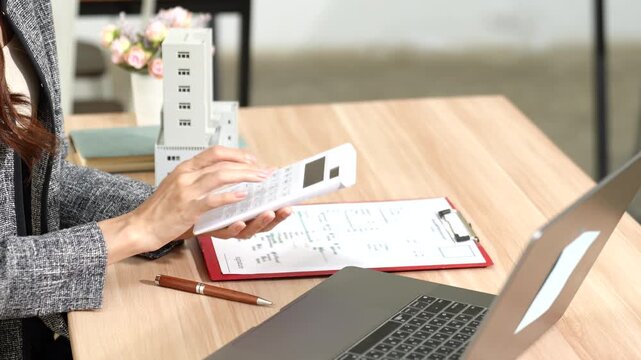 Businesswoman holding a small building model at a desk with a laptop and documents, representing real estate investment, property management, and financial decision-making.