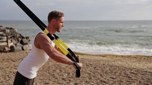 Side view young strong sporty athletic fit sportsman man wear sports clothes use trx loops do push-ups warm up train at sunrise sun dawn over sea beach outdoor on pier seaside in summer day morning