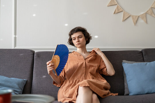 Unmotivated woman coping with summer heat in apartment with no air conditioning using hand fan for cooling, female with sick expression sits on couch staring and waving paper fan, hot season struggles