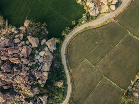 Aerial view of a winding road snakes between rugged, rocky terrain and lush green fields, contrasting natural textures, Hampi, Karnataka, India.