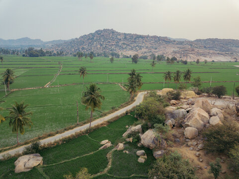 Aerial view of vibrant green paddy fields meet rocky outcrops and winding roads, framed by distant hills, Hampi, Karnataka, India.