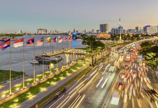 Aerial view of vibrant city lights blurring across the bustling Saigon River promenade, juxtaposed against the calm water and flag displays, Ho Chi Minh City, Vietnam.