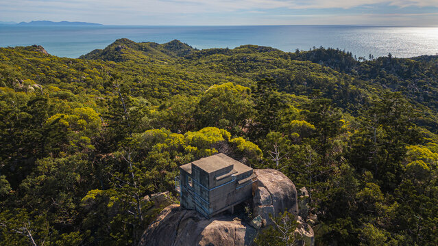 Aerial view of a concrete structure perched atop a rocky outcrop amidst lush green forest, with the vast ocean shimmering in the distance, Magnetic Island, Queensland, Australia.