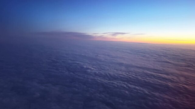 Aerial perspective above clouds at sunrise showing blue sky and a warm orange glow spreading across the cloud layer.