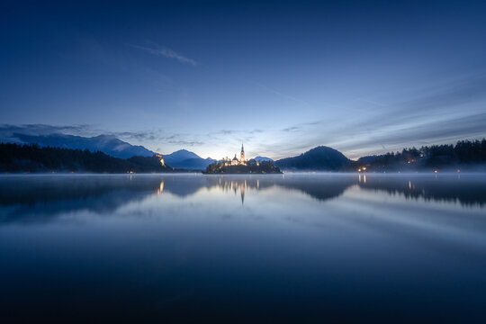 Aerial view of the iconic Bled Island with its church reflected in the tranquil lake under a serene twilight sky, Bled, Radovljica, Slovenia.
