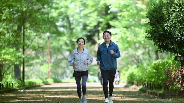 新緑の公園でウォーキングやランニングのスポーツが楽しいミドル夫婦の正面　健康的（ヘルスケア）ライフスタイルのイメージ全身の動画スローモーション