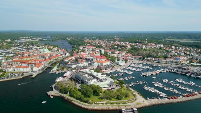 High altitude drone slowly pans across Stromstad revealing full cityscape with orange rooftops, marina packed with yachts, canals, rocky Laholmen peninsula and Scandic hotel