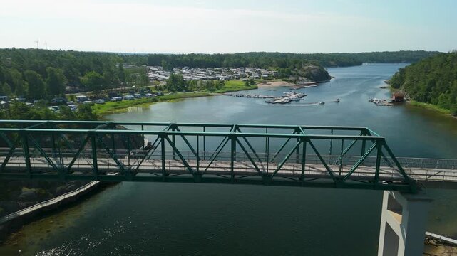 Drone follows vehicle crossing Daft&ouml;bron steel truss bridge with view of holiday camping resort and motorboats in water during summer day in Stromstad Sweden