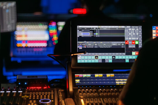 Technicians operate lighting and audio control consoles during a live event, with illuminated screens and a blurred crowd in the background, creating a dynamic concert production atmosphere