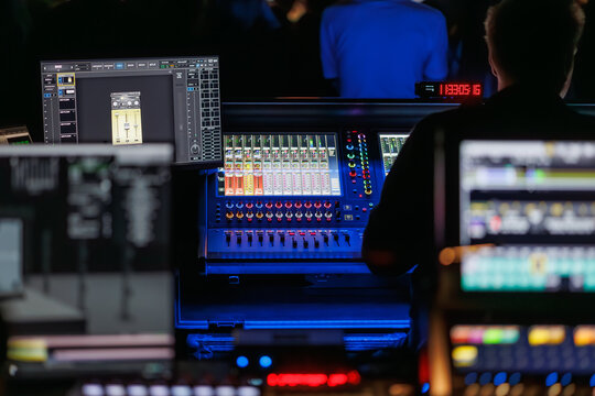 Technicians operate lighting and audio control consoles during a live event, with illuminated screens and a blurred crowd in the background, creating a dynamic concert production atmosphere
