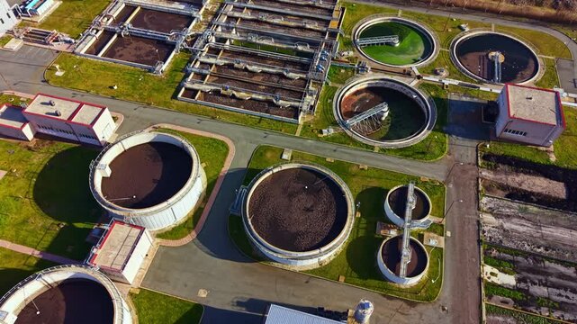 A large wastewater treatment facility features various circular and rectangular tanks along with processing buildings set in an organized layout during daylight.