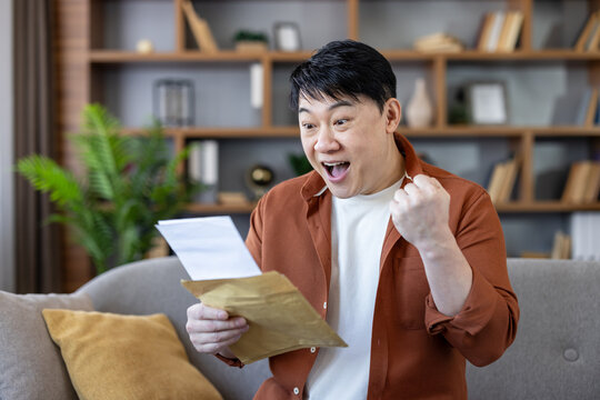 Excited asian man celebrating success while reading a notification letter or document at home, expressing joy and achievement with an open mouth and clenched fist
