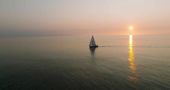 Aerial view of a lone sailboat gliding across the tranquil sea, its white sails contrasting against the golden reflection of the sun, Egmond aan Zee, Netherlands.