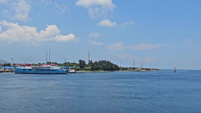Video from a sea vessel approaching Gilimanuk Harbor Bali, showing nearby ships, calm water surface, distant mountains, port structures, and clear sunny sky.