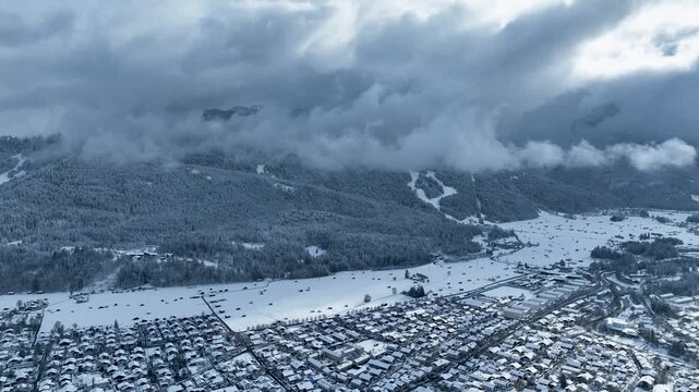 Aerial view of a snow-laden landscape with a town nestled against a backdrop of mountains shrouded in clouds, Garmisch-Partenkirchen, Bavaria, Germany.