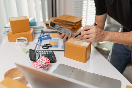 Small business owner checking order on smartphone while writing on parcel box, preparing packages for delivery, managing ecommerce workflow and shipping process on organized workspace.